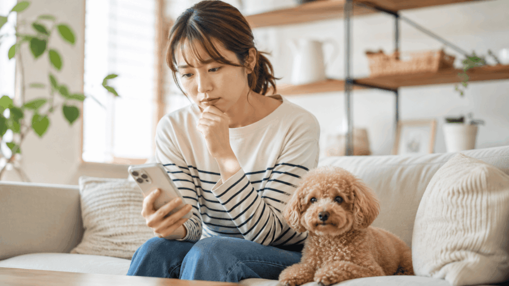 トリミングサロンの予約について悩みながらスマートフォンを見る飼い主さん。隣には少し毛が伸びてきた小型犬が寄り添っており、“予約が取れない…”という現代の愛犬家の悩みを表現した日常風景。