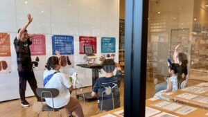 講師と参加者が挙手しながら対話している様子。ガラス越しにイベントの活気が伝わる「イヌの学び舎」の一場面。