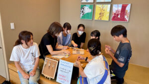 室内のワークショップで、子どもたちと大人がウッドビーズのペアチャームづくりに挑戦している様子。