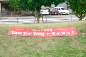 芝生の上に広げられた赤い横断幕。「犬と人の運動会 One for Dog トライアル！」の文字が印刷されている。背景には木と道路が見える。
