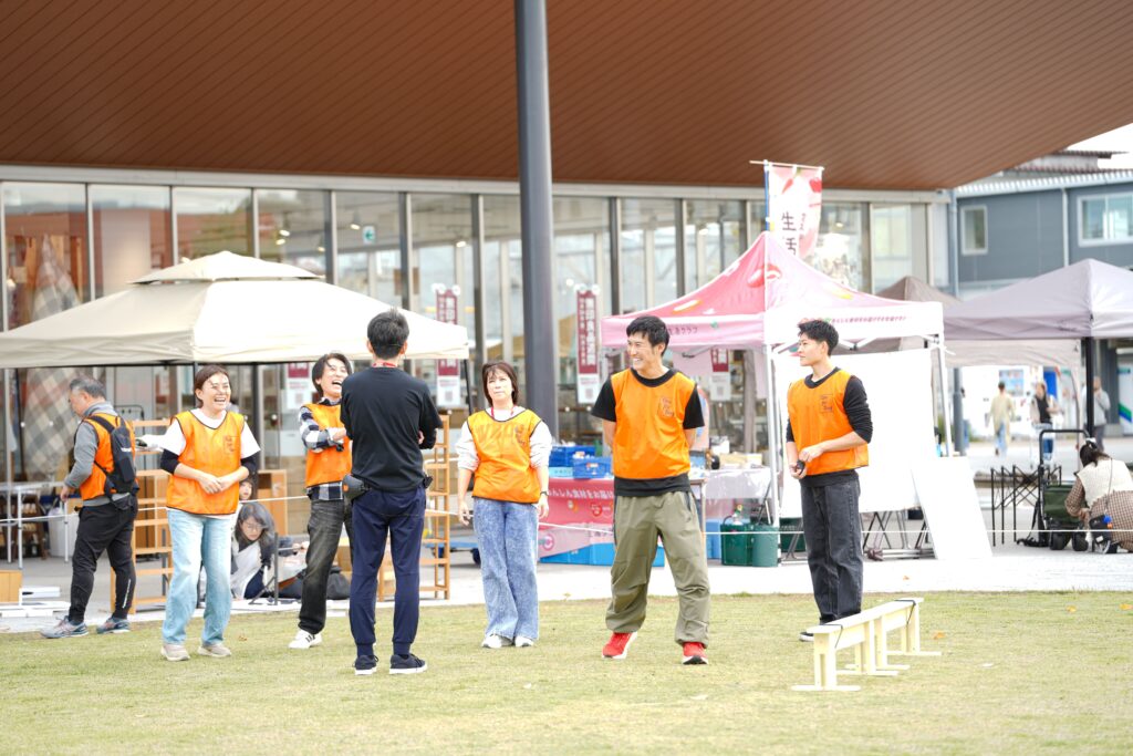 イベント会場の前で、オレンジのビブスを着たスタッフたちが打ち合わせをして笑顔を見せている。背景には無印良品の店舗とテントブース。