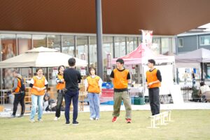 イベント会場の前で、オレンジのビブスを着たスタッフたちが打ち合わせをして笑顔を見せている。背景には無印良品の店舗とテントブース。