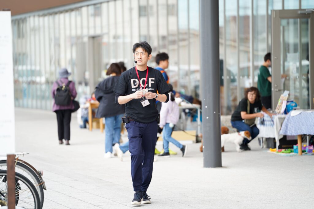 黒いTシャツに「DCF」のロゴを付けたスタッフがマイクを装着して会場内を歩いている。背後には来場者と犬たちの姿が見える。