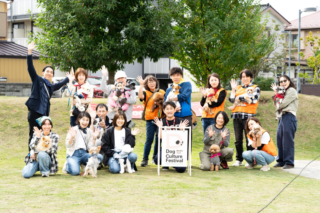 「Dog Culture Festival」の看板を中心に、スタッフやボランティア、飼い主たちが愛犬を抱いて記念撮影。全員が笑顔で手を振っている集合写真。