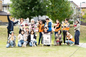 「Dog Culture Festival」の看板を中心に、スタッフやボランティア、飼い主たちが愛犬を抱いて記念撮影。全員が笑顔で手を振っている集合写真。