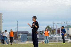 屋外の芝生広場で、マイクを付けた進行スタッフが参加者に向けて話している。背後にはオレンジのビブスを着たスタッフや犬を連れた来場者の姿。