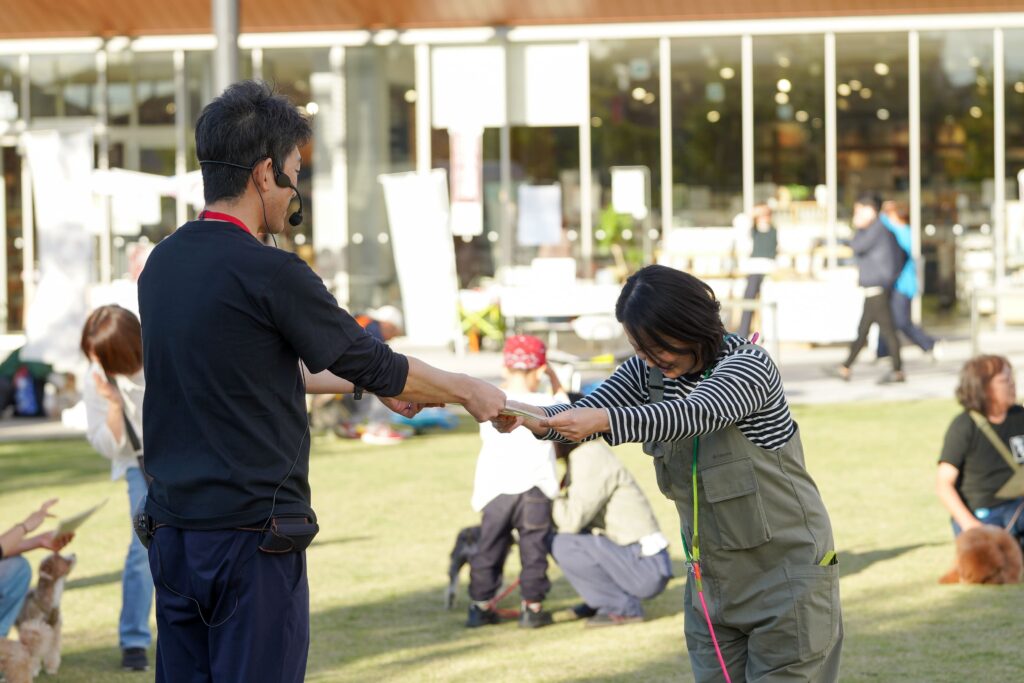 屋外会場で、進行スタッフが女性参加者に賞状を手渡している。2人が笑顔でお辞儀を交わし、周囲には犬たちと観覧者の姿が見える。