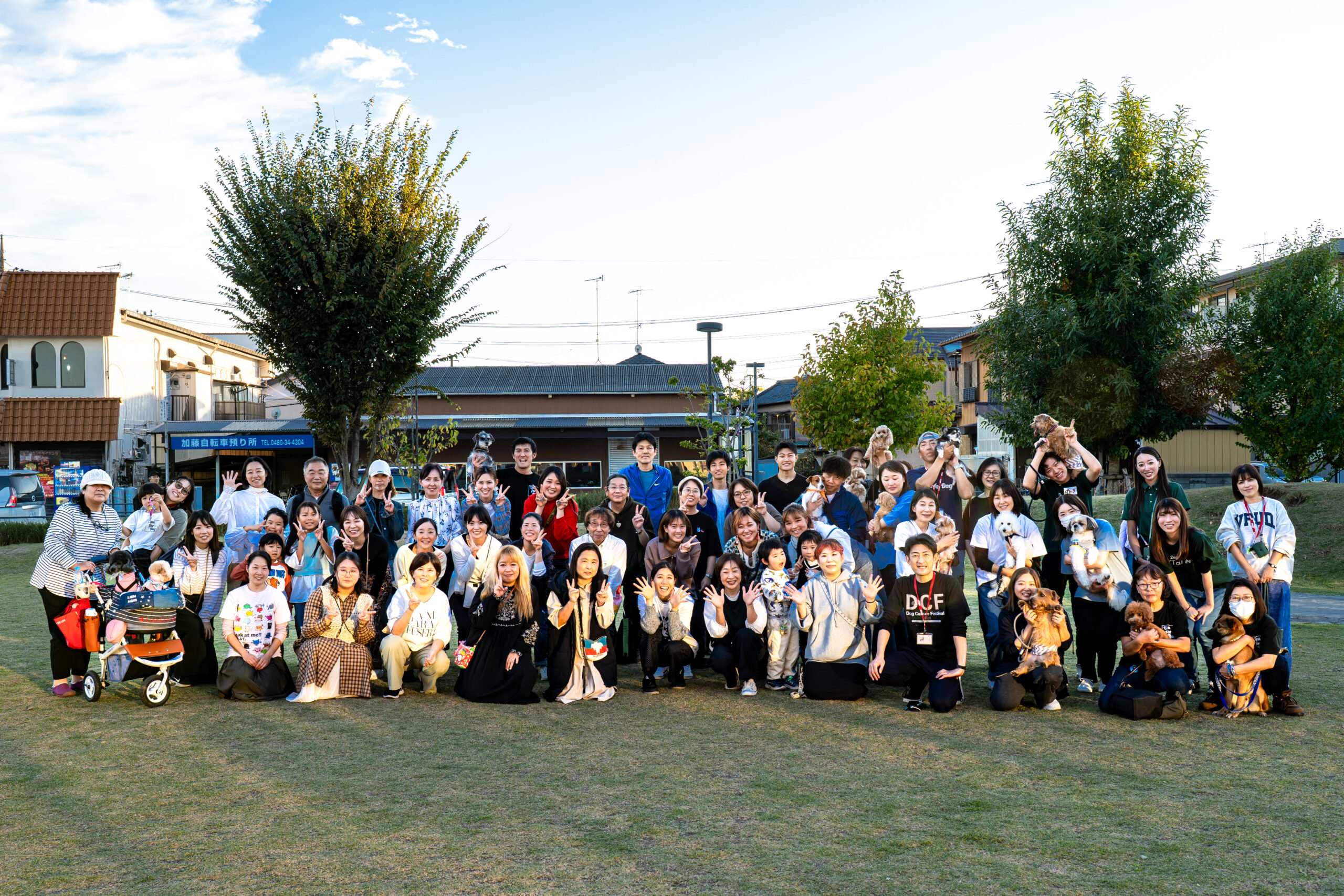夕方の芝生広場で、愛犬と飼い主、スタッフが一堂に集まった大集合写真。笑顔でピースサインをする人や犬を抱く人など、フェスの締めくくりを飾る記念撮影の様子。