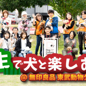 芝生の広場で、愛犬と一緒にポーズをとる参加者たちの集合写真。背景には木々と施設があり、前面には『芝生で犬と楽しむ会@無印良品 東武動物公園駅前』の大きなタイトルロゴが配置されている。