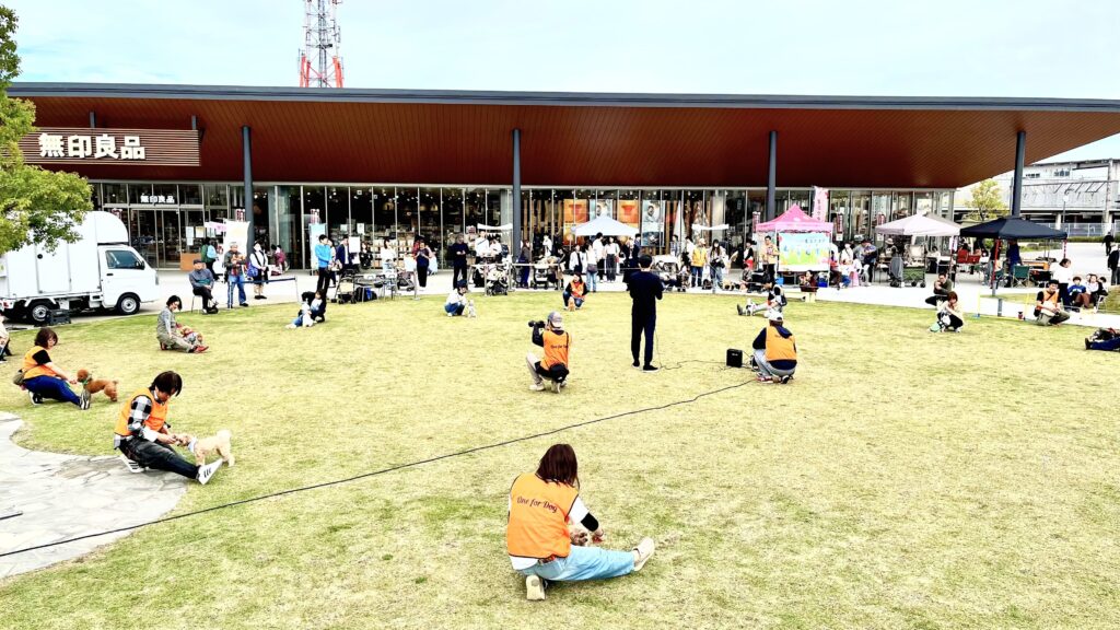 無印良品 東武動物公園駅前の芝生広場で開催されたイベントで、飼い主と愛犬が一緒にドッグダンスをしている様子。One for Dogのスタッフが指導を行い、来場者が見守る光景。