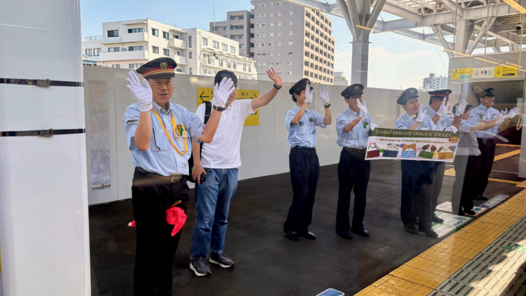 駅のホームで、TOBU Doggy Doggy Trainの横断幕を持ち、列車に手を振る東武鉄道のスタッフ