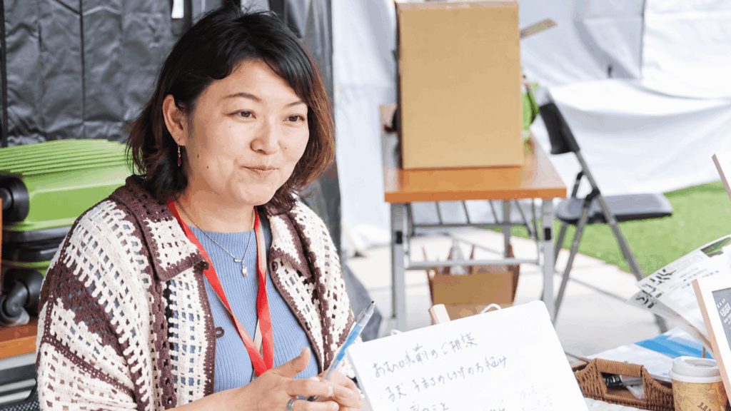 屋内のイベント会場で、赤いストラップと白・茶色の編み柄のカーディガンを身につけた女性が、ペンを手に穏やかな表情で座っている。手前には手書きの案内板、背景には椅子や段ボール箱が見える。